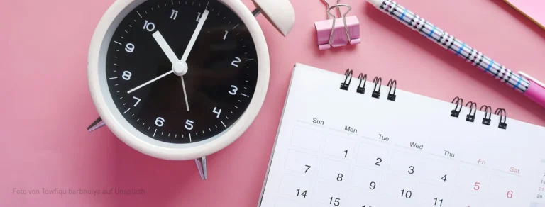 Planung & Timing: Wann sollte man Kalender drucken? Ein Wecker neben einem aufgeschlagenen Tischkalender neben Stiften auf rosa Untergrund.