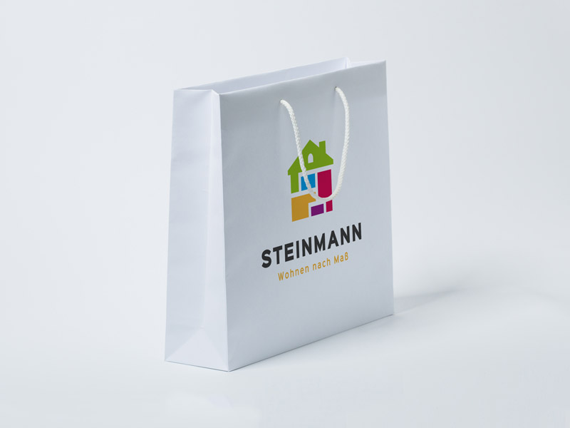 Die Abbildung zeigt eine weiße Papiertasche im Querformat aus einerseitlichen Ansicht. Sie hat eine weiße Stoffkordel und ein buntes Logo als Aufdruck.