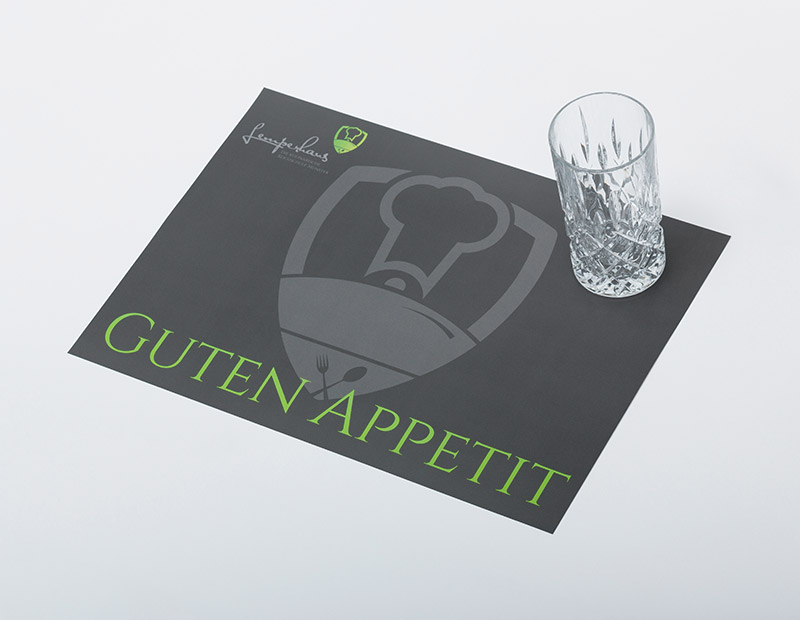 Ein dunkelgrau mit grüner Aufschrift gehaltenes Tischset, worauf oben rechts am Bildrand ein Wasserglas positioniert ist